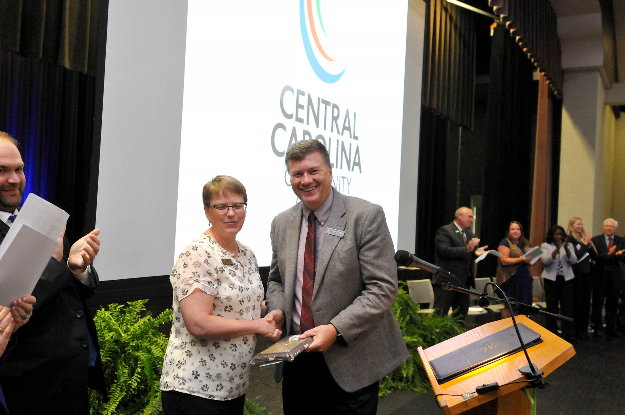 Click to enlarge,  Central Carolina Community College Dean of Arts, Sciences and Advising Scott Byington (right) receives the Staff Member of the Year award from 2018 recipient and CCCC Student Learning Coordinator Angela Crisp-Sears (left) during the Convocation and Installation of Dr. Lisa M. Chapman as Sixth President of CCCC on Sept. 26. 
