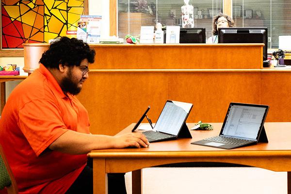 Student sitting at library table interacting with a few different electronic devices  